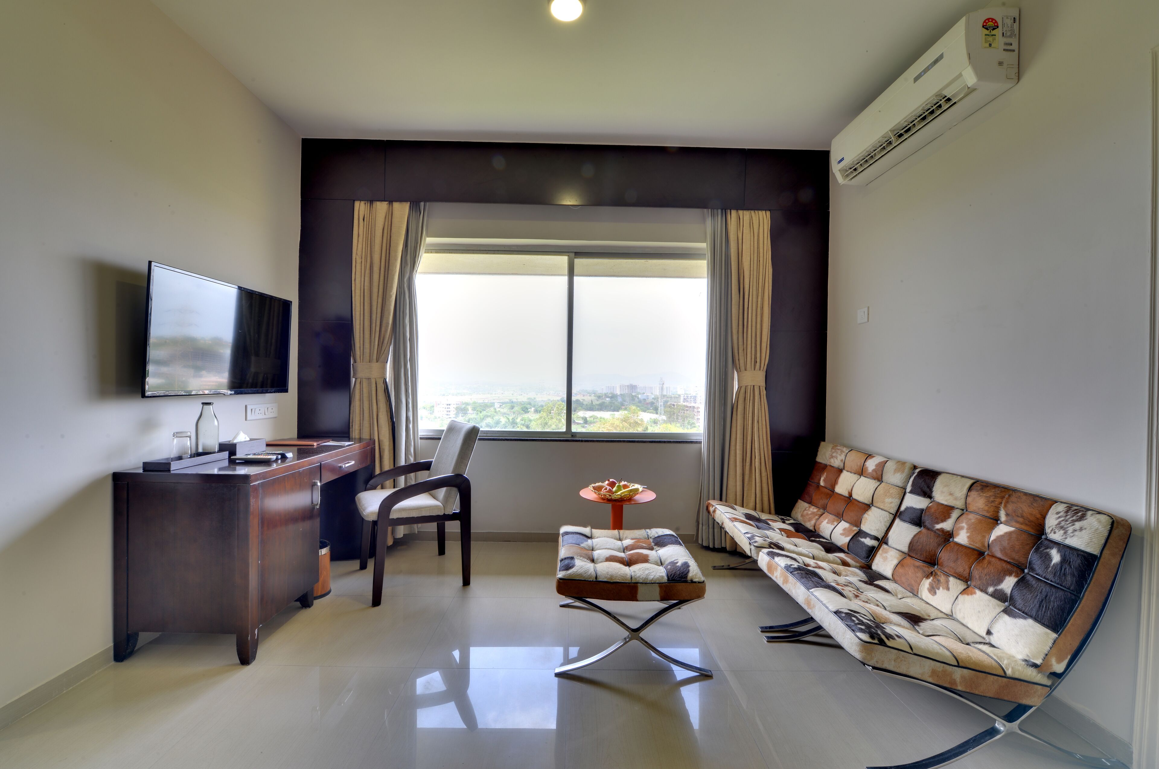 A modern living room with a flat-screen TV, a wooden desk, a cushioned stool, a sofa, and a large window with curtains.