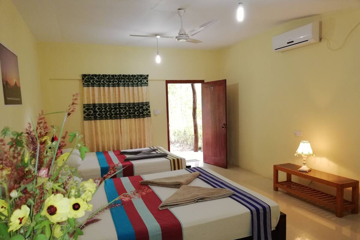 A hotel room with two beds, a ceiling fan, an air conditioner, a wooden nightstand with a lamp, and a window with curtains.