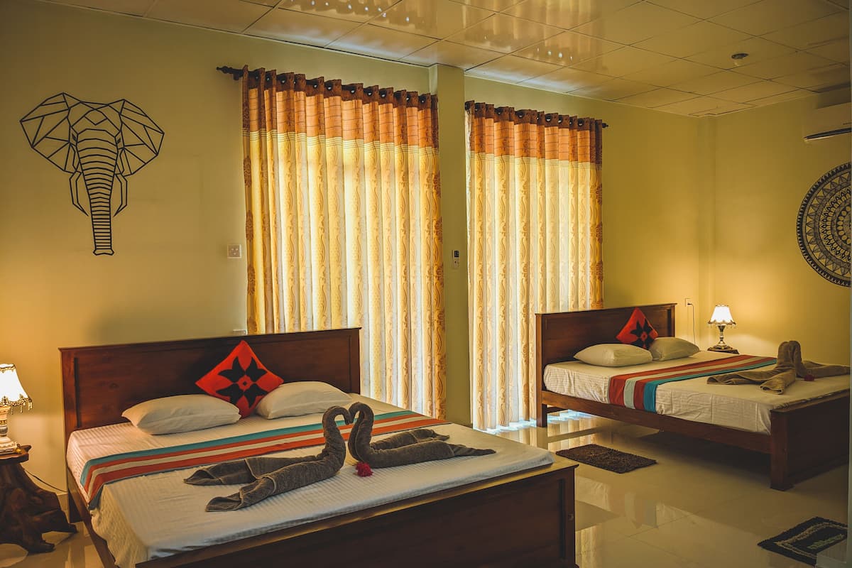 A hotel room with two beds, each with a wooden headboard, a red and black pillow, and a swan towel arrangement. There are curtains with a patterned design, a wall tapestry, and a round wall decoration.