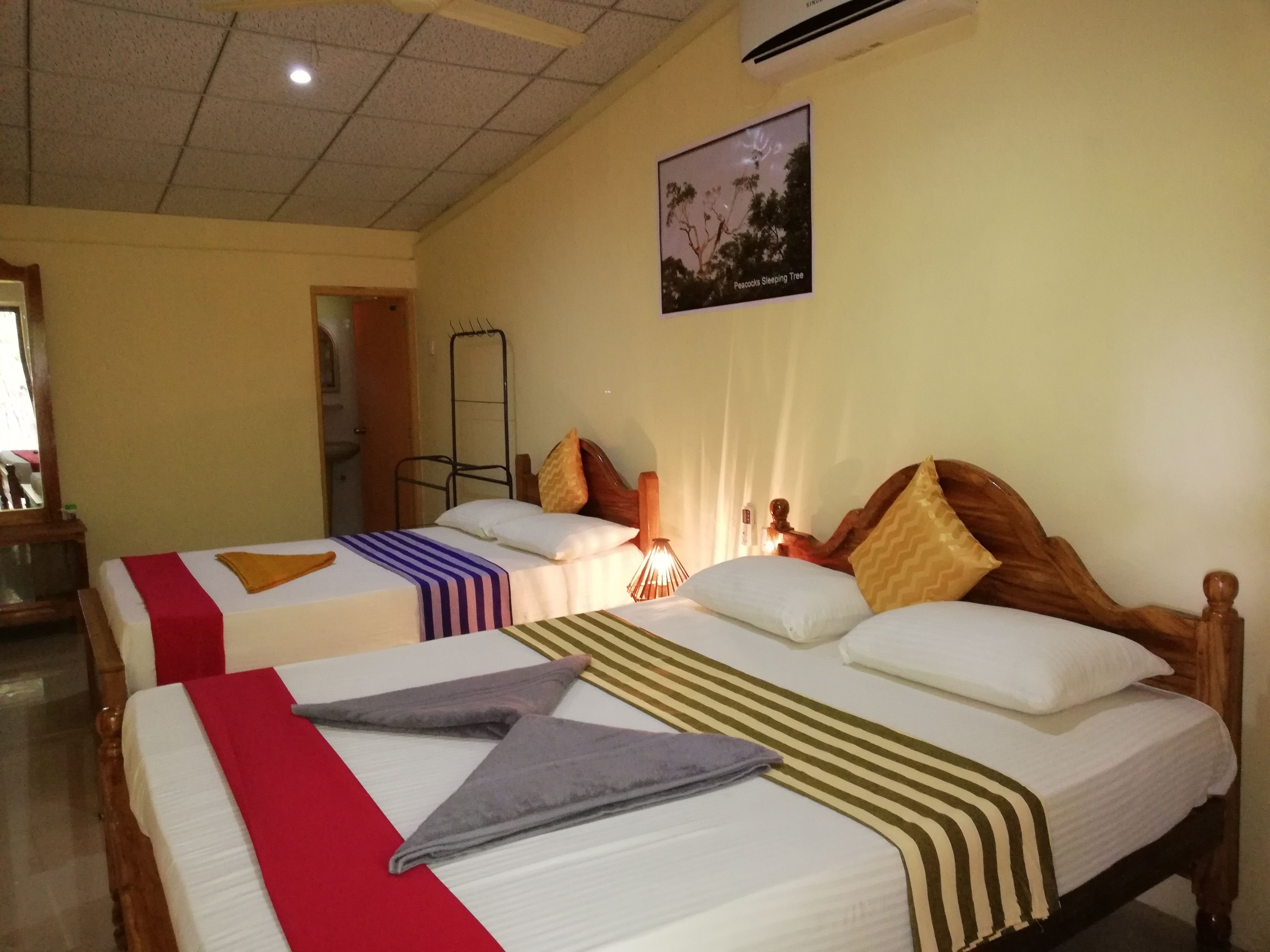 Two single beds with headboards, a wall-mounted air conditioner, a framed picture of a tree, and a mirror on the left wall.
