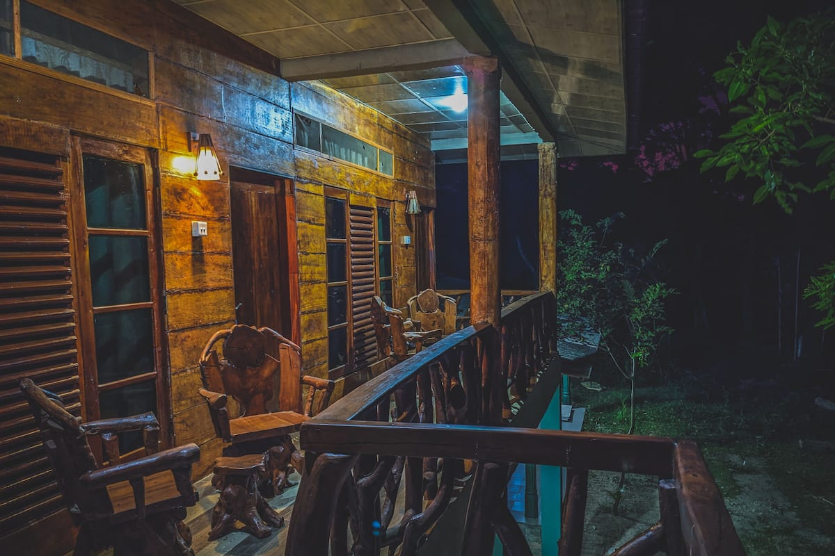 A rustic wooden porch with a wooden bench and chairs, a railing, and a view of a garden.