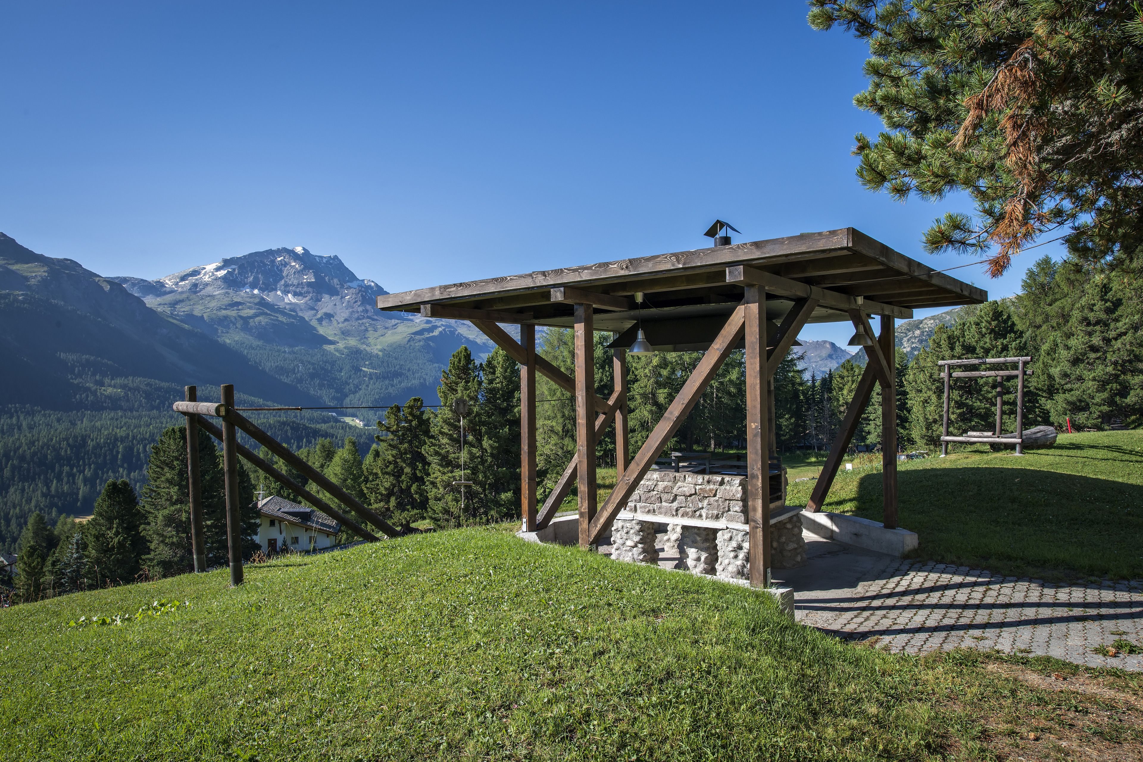 Ein hölzerner Pavillon mit Steinstufen, gelegen auf einem grasbewachsen Hügel vor einer Bergkulisse.