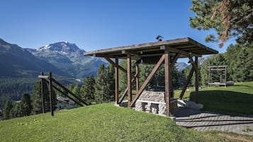 Ein hölzerner Pavillon mit Steinstufen, gelegen auf einem grasbewachsen Hügel vor einer Bergkulisse.