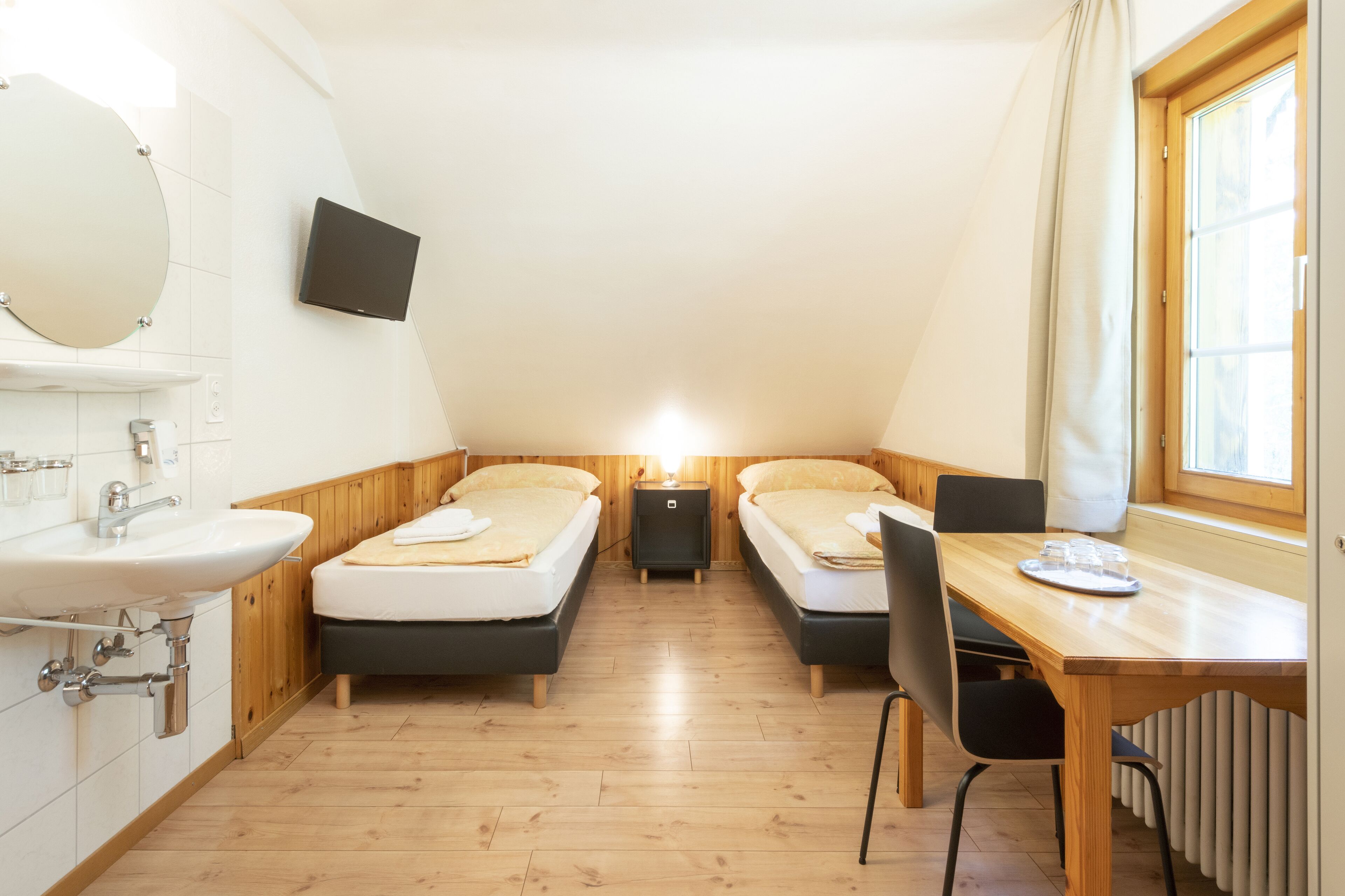 A small, compact room with two beds, a wooden table, a sink, and a TV mounted on the wall.