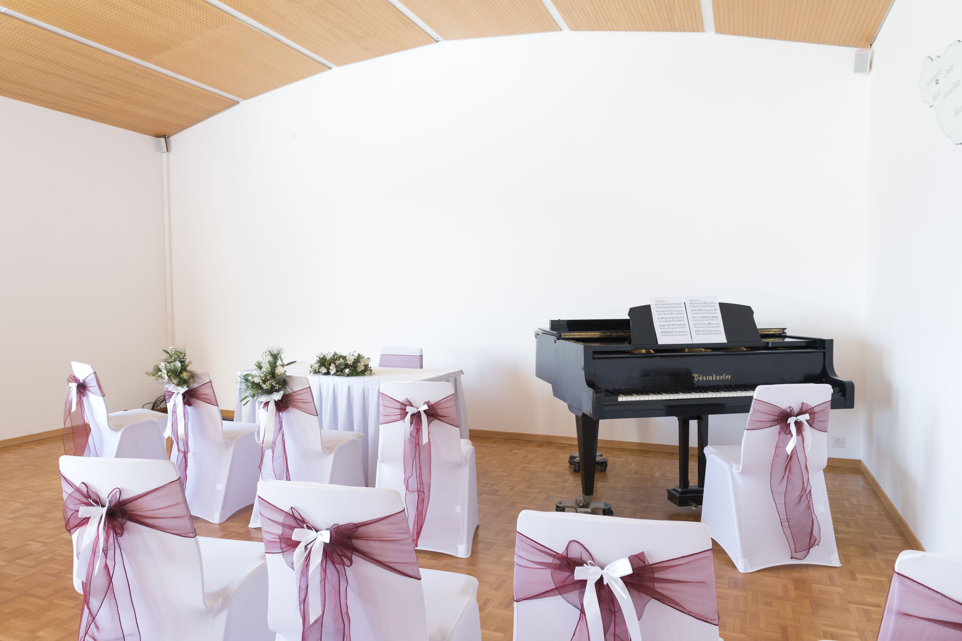 Ein Zimmer mit einem Flügel, weißen Stühlen mit rosa Schleifen und einem weißen Tisch mit Blumenarrangement.