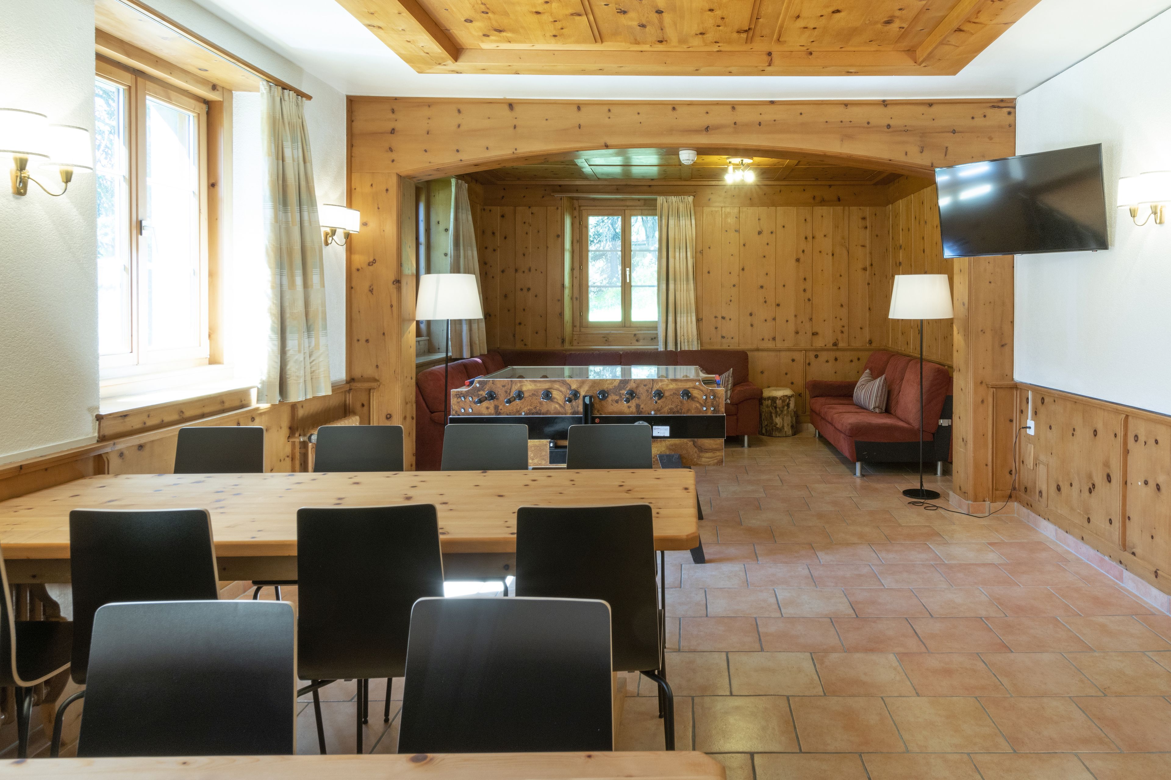 A wooden-paneled room with a large table and chairs, a sofa, and a television mounted on the wall.