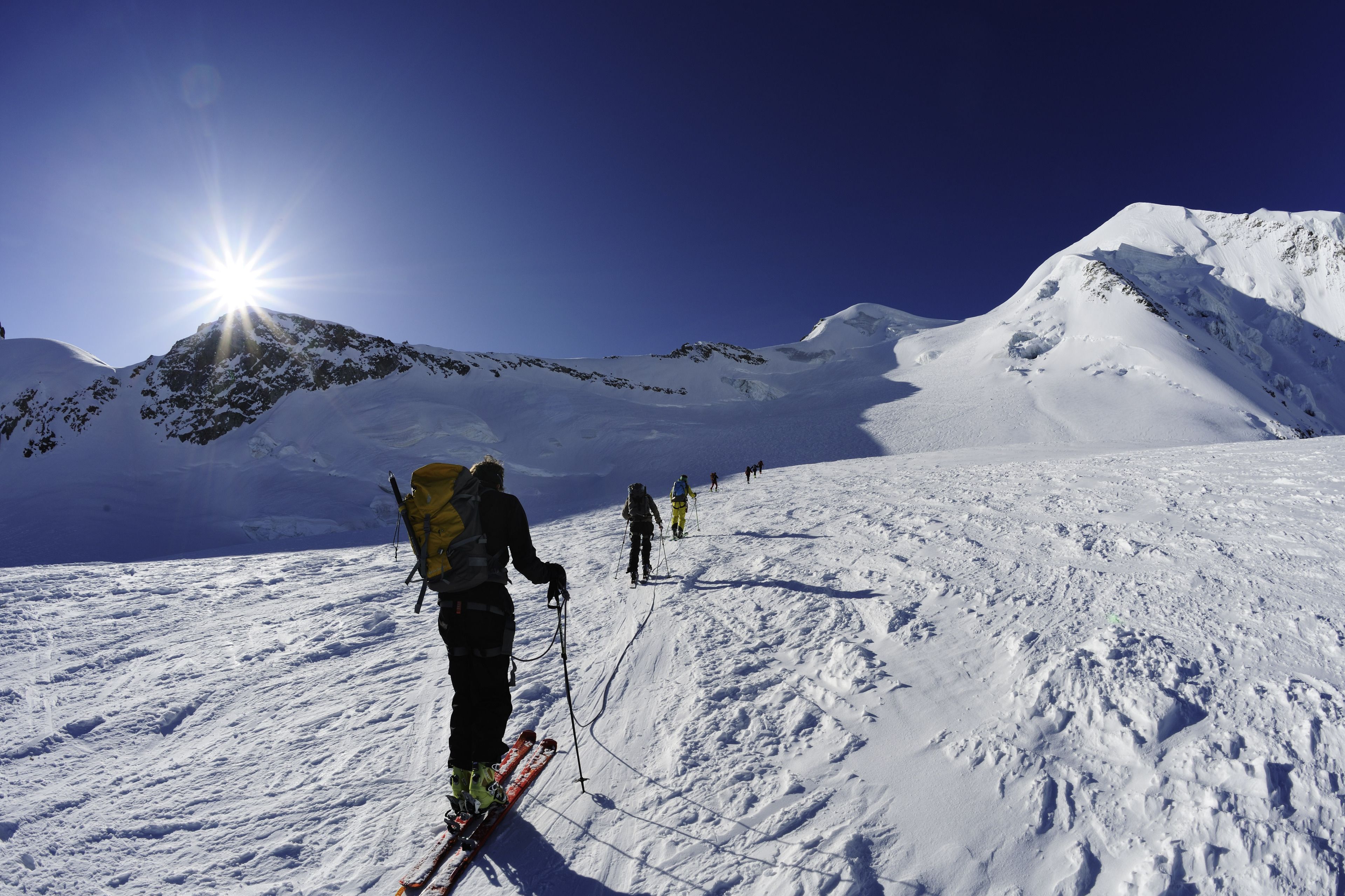 Paesaggio montuoso innevato con sciatori che salgono una pista sotto un cielo sereno.