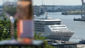 Un grand navire de croisière était amarré à un port, avec des bâtiments industriels et des éoliennes en arrière-plan.