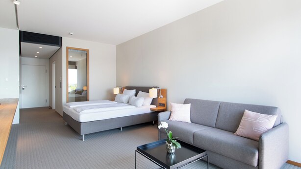 Une chambre d’hôtel moderne avec un lit double, un canapé gris, une petite table avec un vase et un miroir au mur.