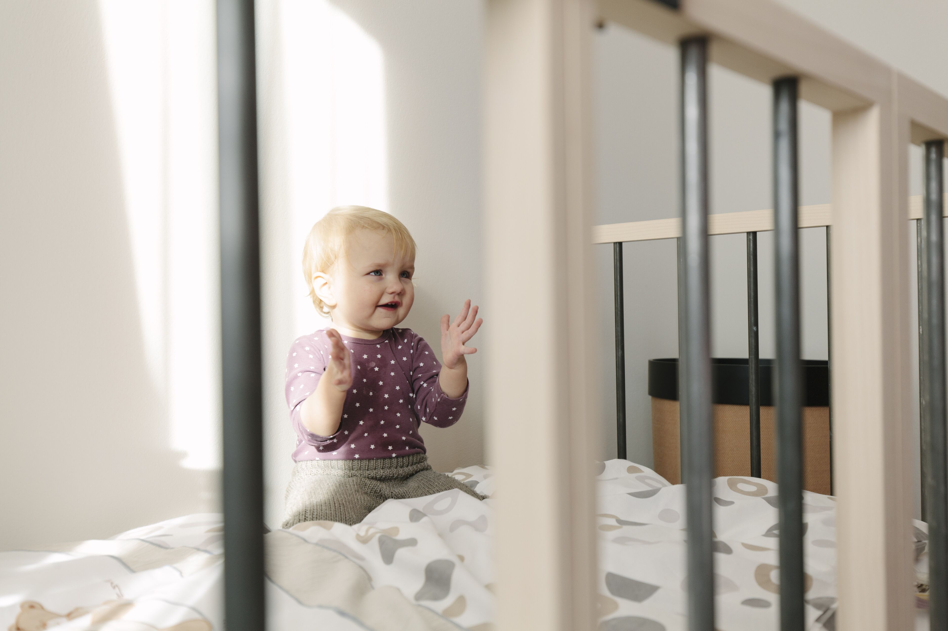 Niño sentado en una cama con colcha estampada, una cuna al fondo y una ventana que deja entrar luz natural.