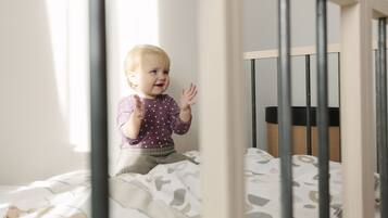 Niño sentado en una cama con colcha estampada, una cuna al fondo y una ventana que deja entrar luz natural.