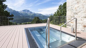 Piscina rectangular al aire libre con barandillas metálicas, rodeada por una cubierta de madera y un muro de piedra, con vistas a un paisaje montañoso.
