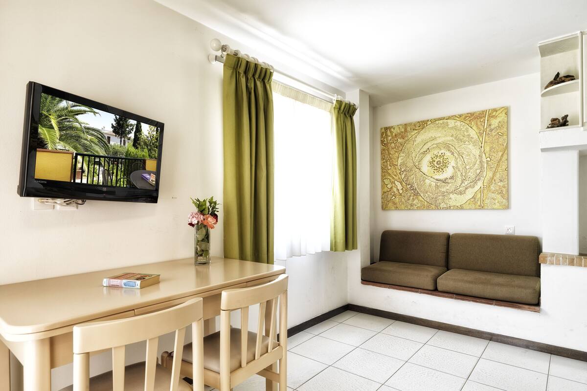 Ein Wohnzimmer mit Sofa, einem Wandfernseher, einem Tisch mit Stühlen, einem Fenster mit Vorhängen und einem Wandbild.