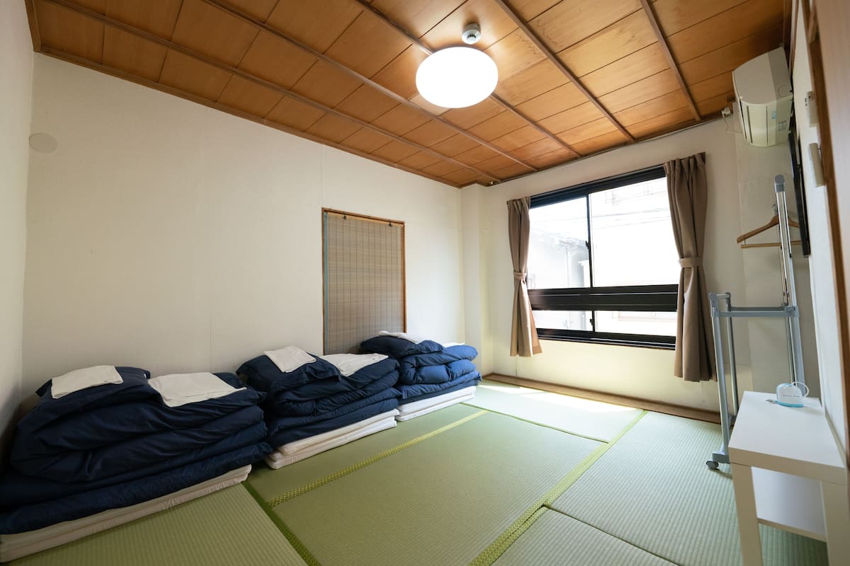 A room with tatami mats, a wooden ceiling, a bed with blue bedding, a window with curtains, and a small table with a cup.