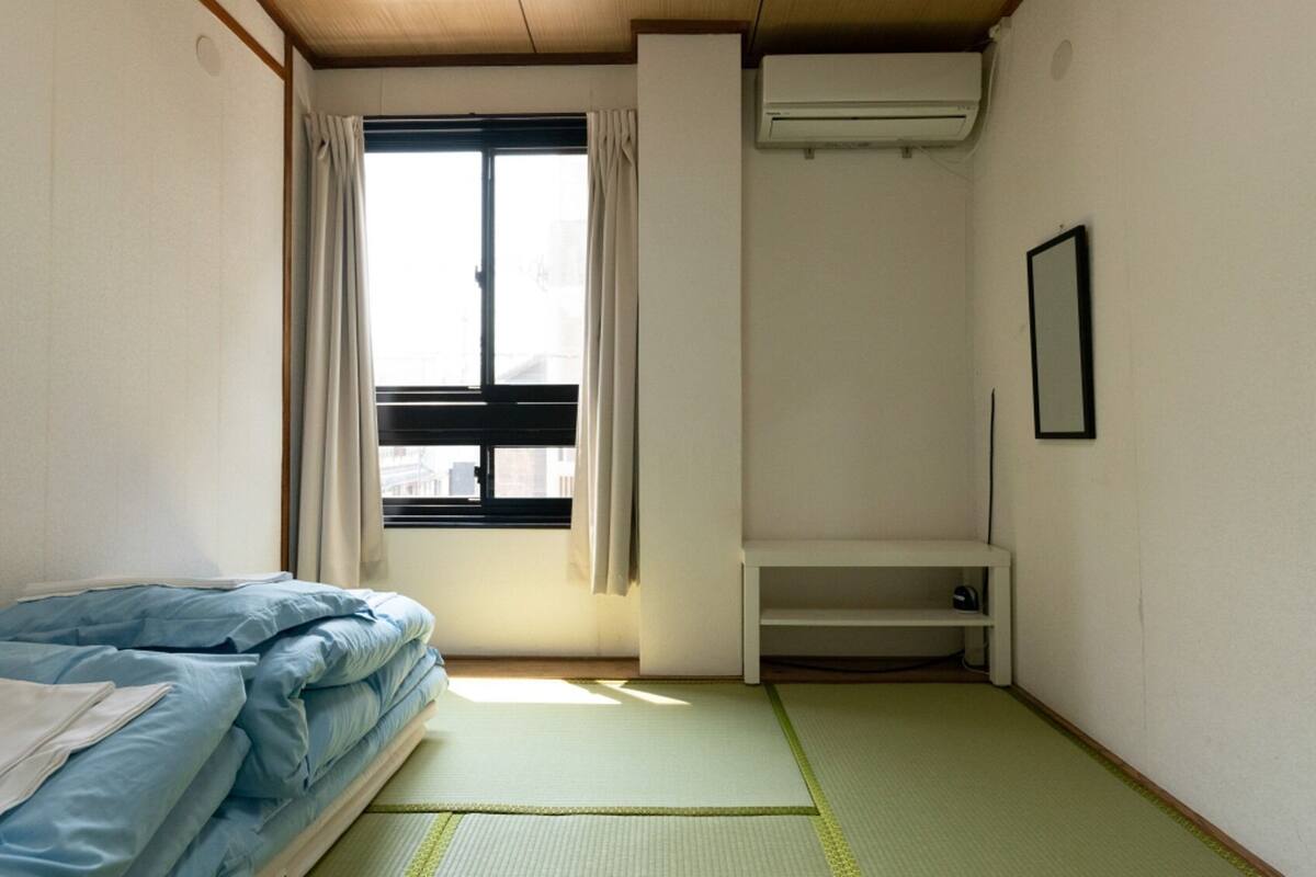 A small, sparsely furnished room with a bed, a window with curtains, a shelf, and an air conditioning unit.