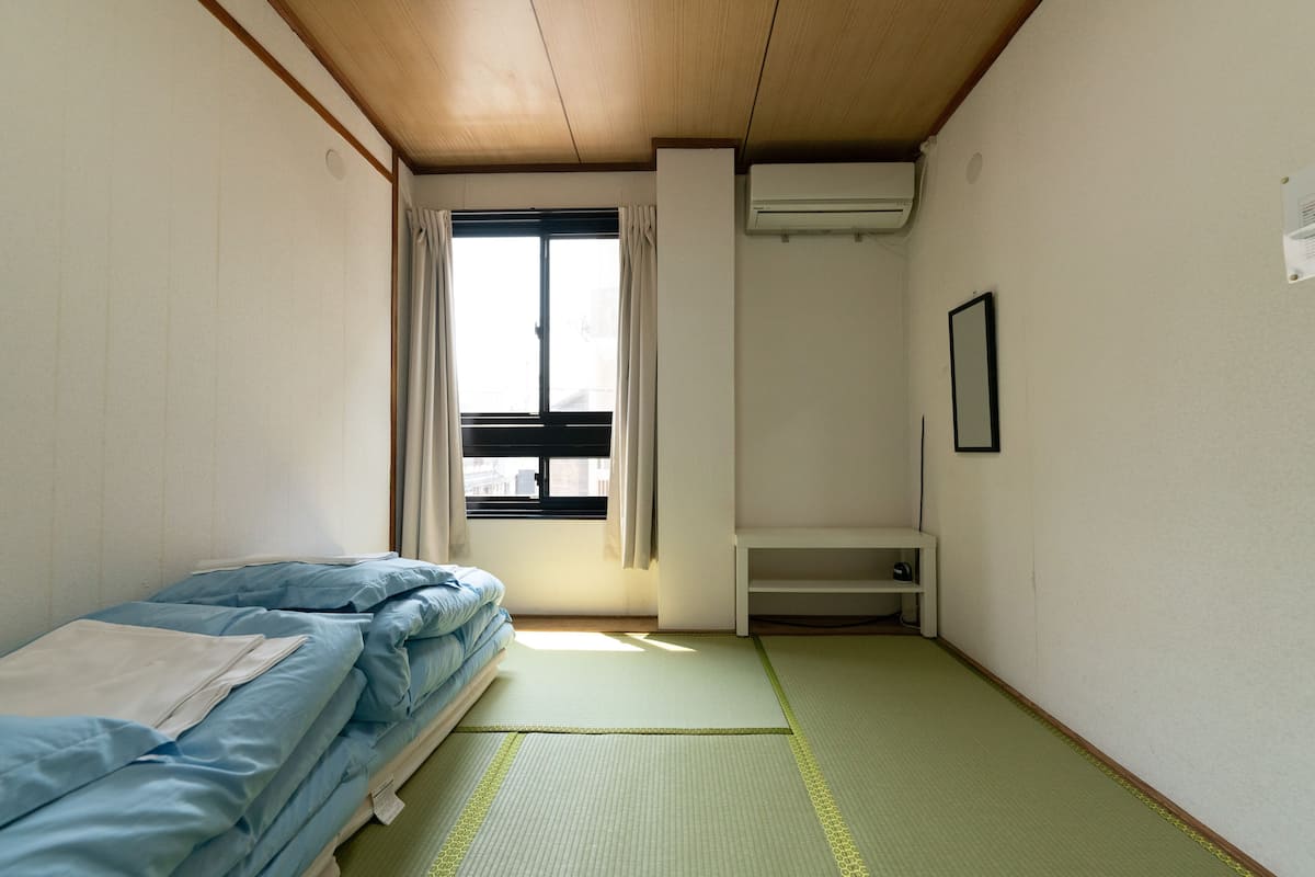 A small, sparsely furnished room with a bed, a window with curtains, a white shelf, and a wall-mounted air conditioner.