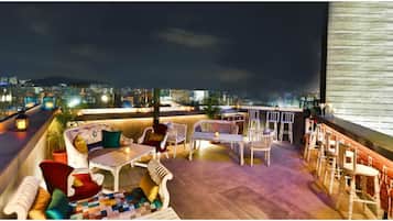 Terraço no telhado com área de estar, bar e vista noturna da cidade.