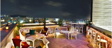 Terraza en la azotea con mobiliario, zona de bar y vista nocturna de la ciudad.