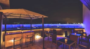 Una terraza al aire libre con mesas y sillas, un toldo y vistas de la ciudad iluminada de noche.