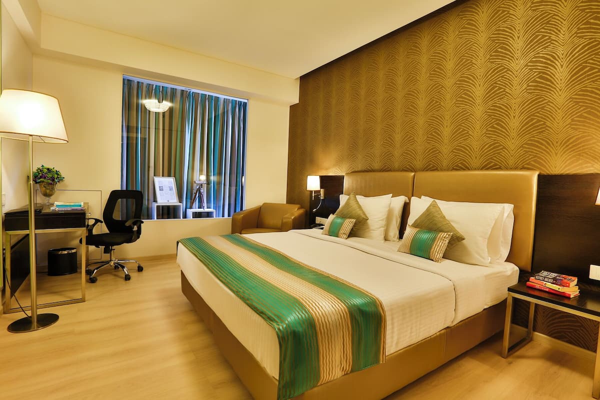 A hotel room with a large bed, a desk with a chair, a lamp, and a window with curtains.