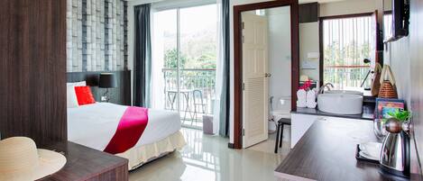 A hotel room with a bed, a desk, a bathroom, and a balcony with a view of trees.