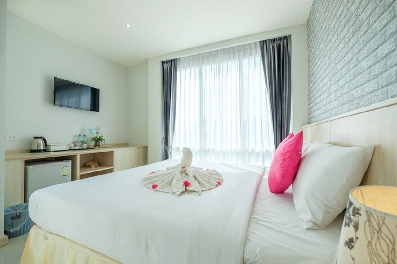 Une chambre d’hôtel avec un lit bien fait, un téléviseur à écran plat fixé au mur, un petit bureau avec une bouilloire et une plante, et une fenêtre avec des rideaux légers.