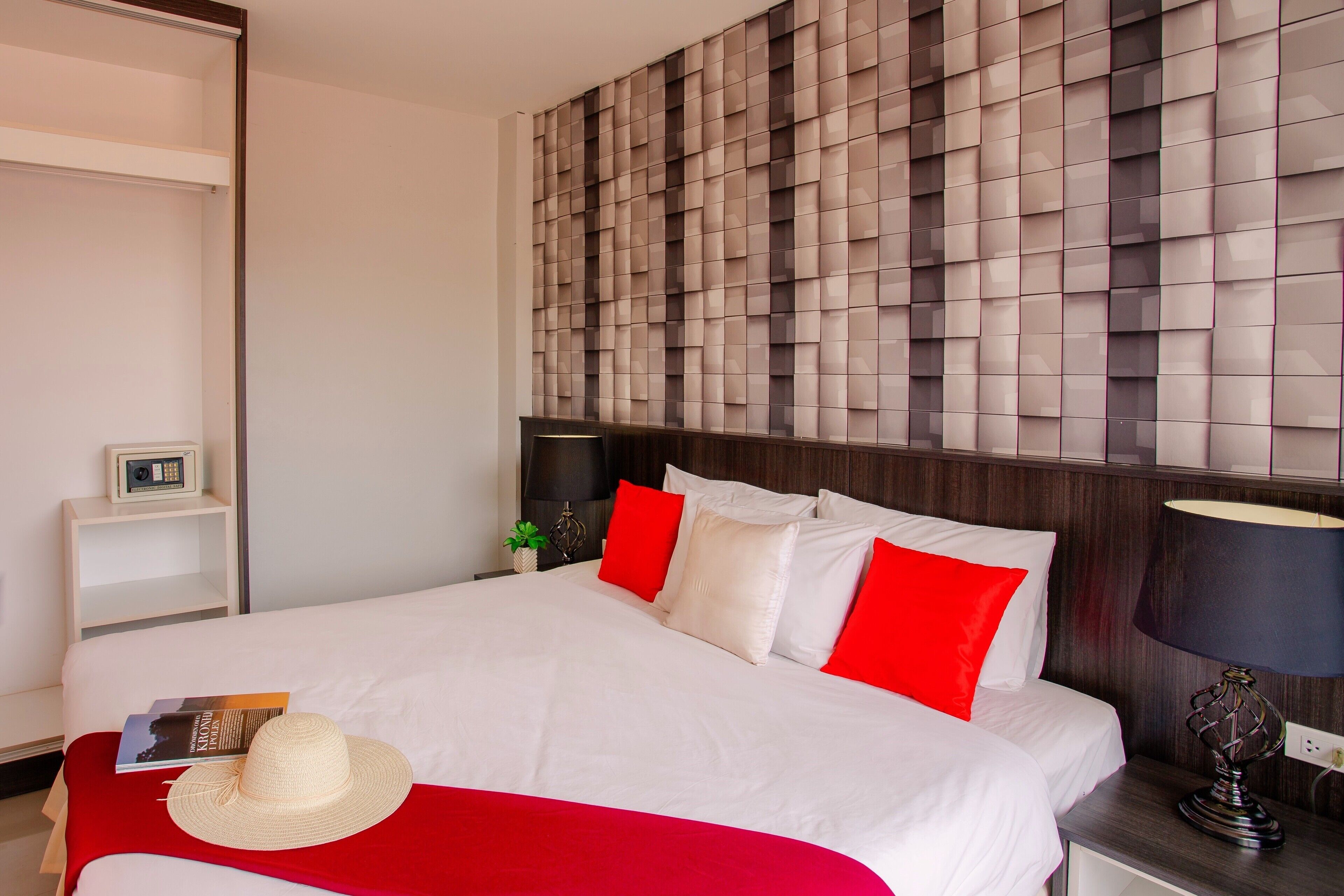 A hotel room with a large bed, red and white bedding, a bedside table with a lamp, and a wall panel.