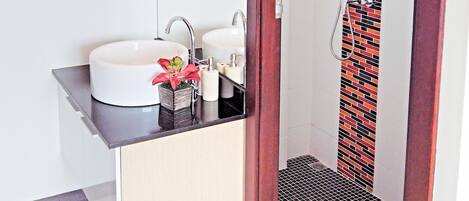 A bathroom with a white sink, two faucets, a mirror, a shower with a patterned curtain, and a small potted plant.