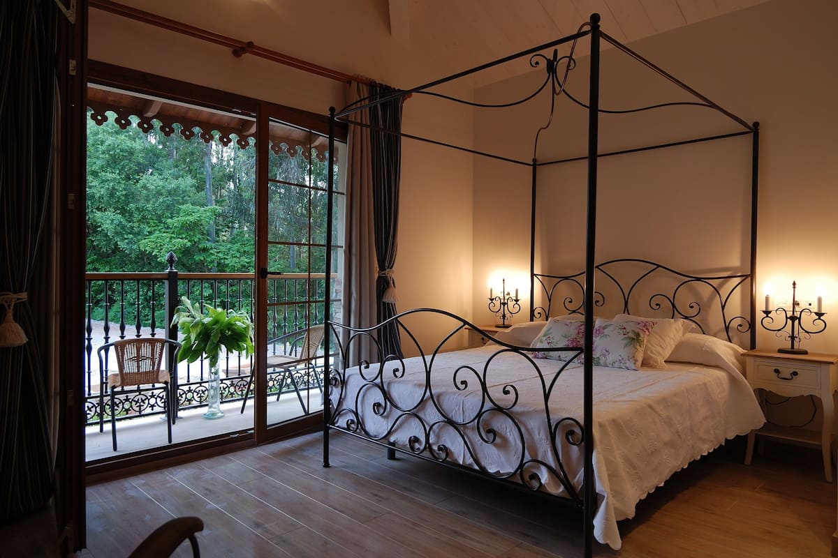 A four-poster bed with a white bedspread, two bedside tables with candelabras, and a view of a balcony with a chair and potted plant.