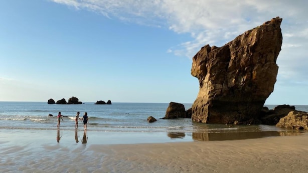 A beach with a large rock formation and three people walking on the sand.