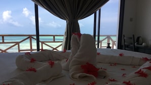 A bed with white towels and red flowers, a view of the ocean through a balcony railing, and a window with curtains.