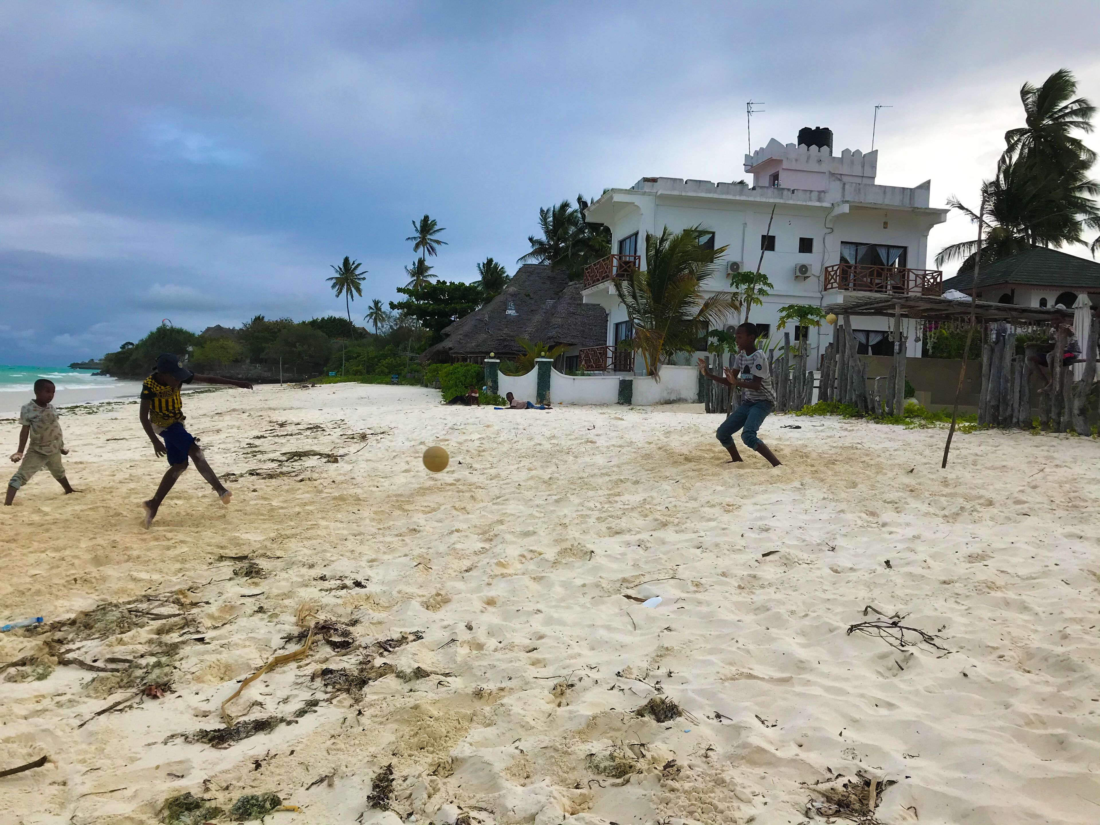 A sandy beach with palm trees and a white building with a thatched roof in the background.