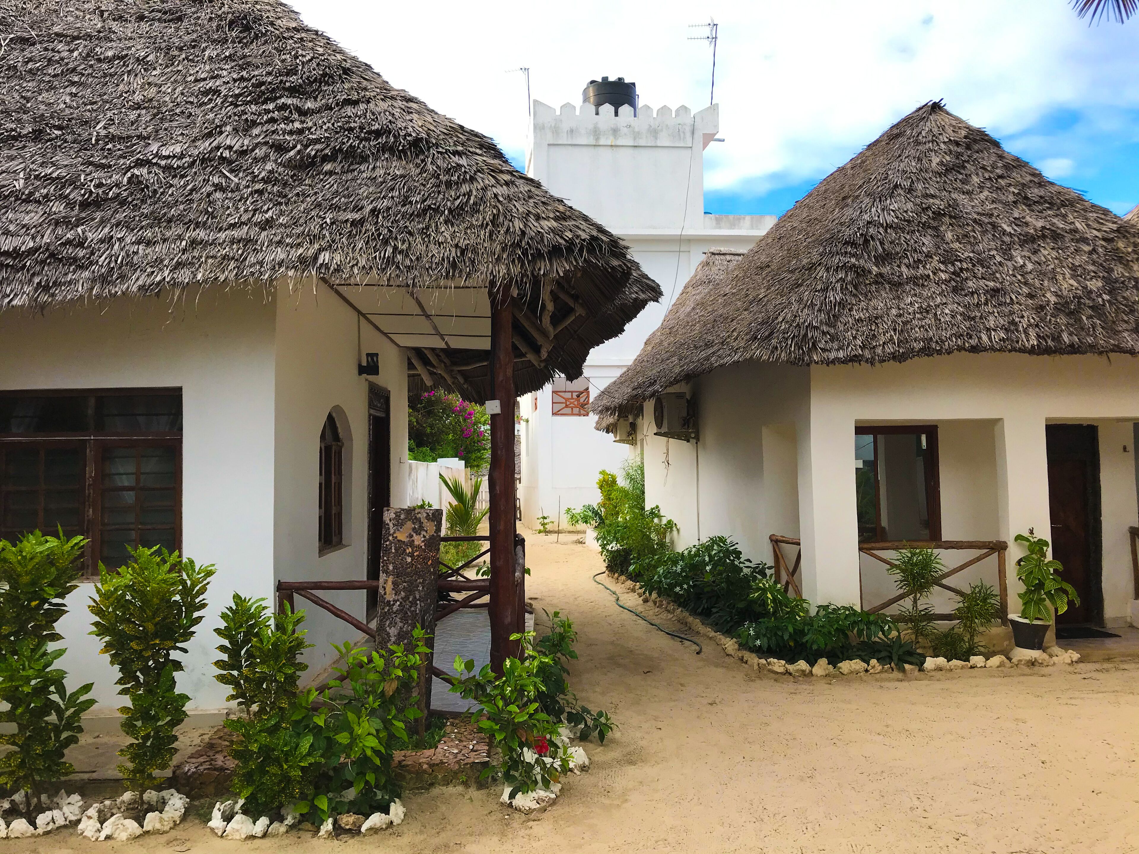 A pathway leading to thatched-roof cottages with white walls, surrounded by greenery and a clear sky.