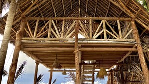 A thatched-roof hut with a wooden framework, surrounded by palm trees, overlooking a beach.