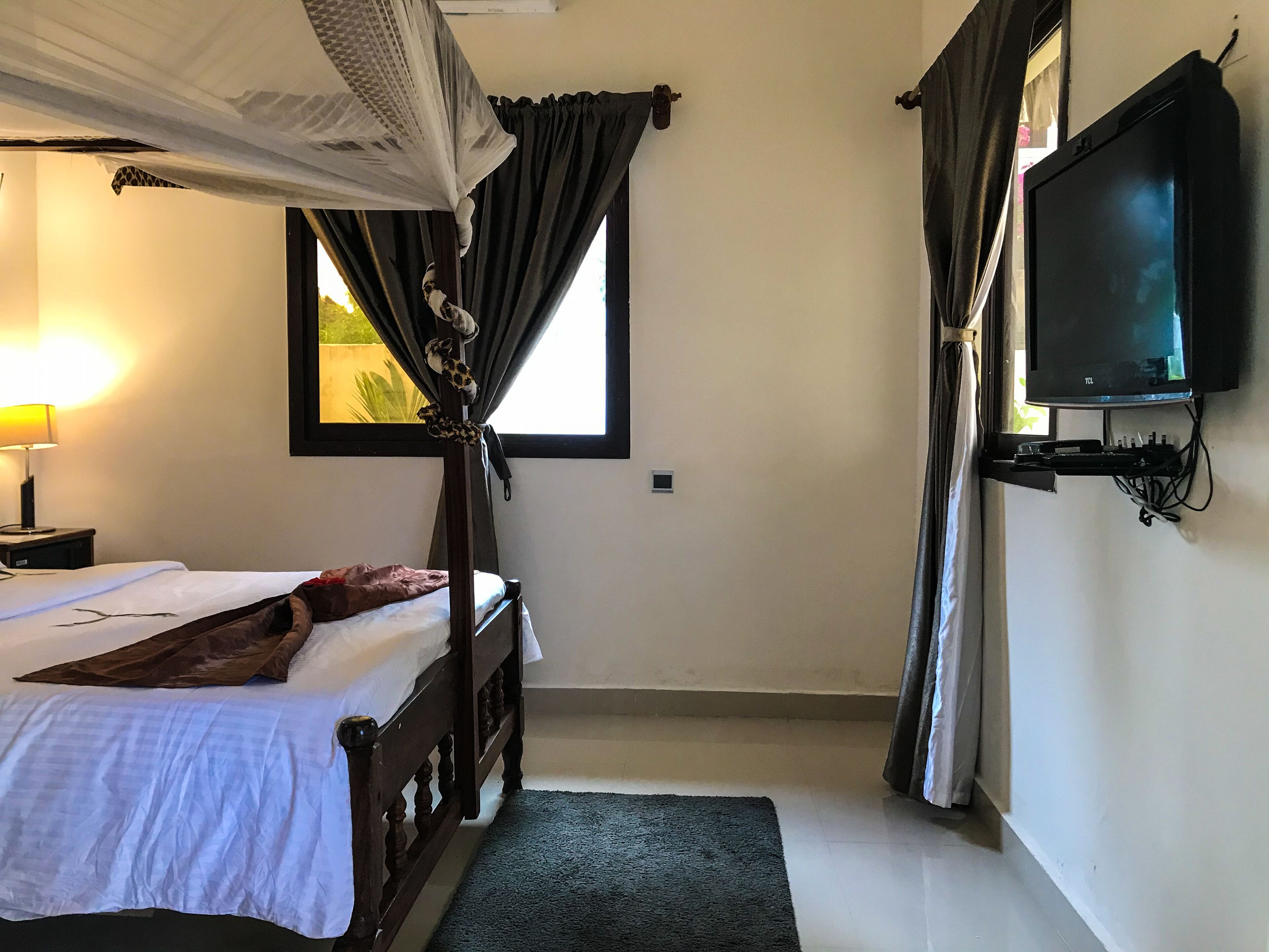 Ein Schlafzimmer mit einem Bett, einem an der Wand montierten Fernseher, einem Fenster mit Vorhängen und einer Nachttischlampe.