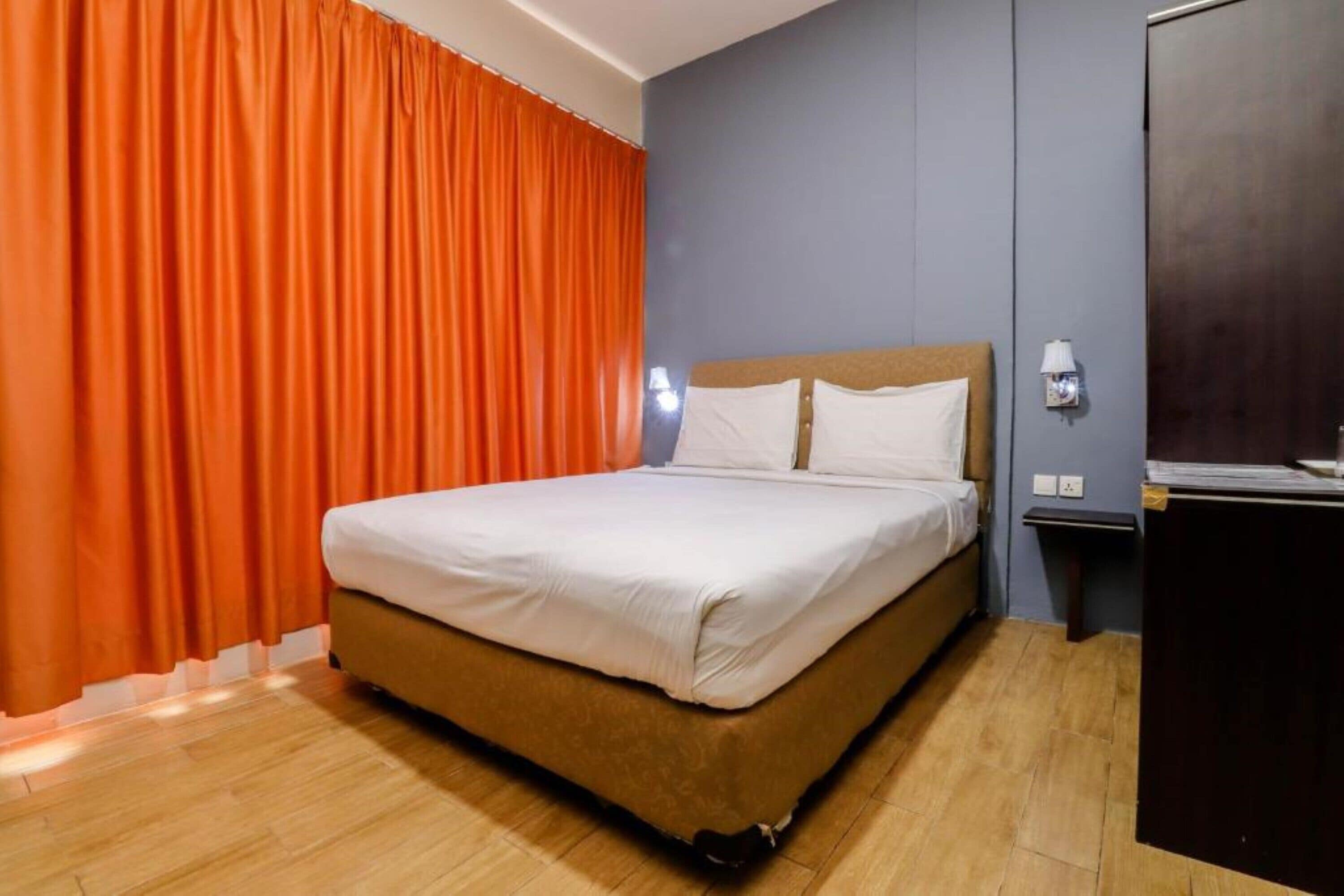 Een hotelkamer met een bed, oranje gordijnen, een nachtkastje en een aan de muur bevestigde lamp.