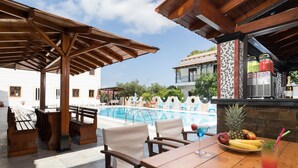 Zona junto a la piscina con asientos de madera, una mesa con fruta y una bebida, y un bar con fruta y bebidas.