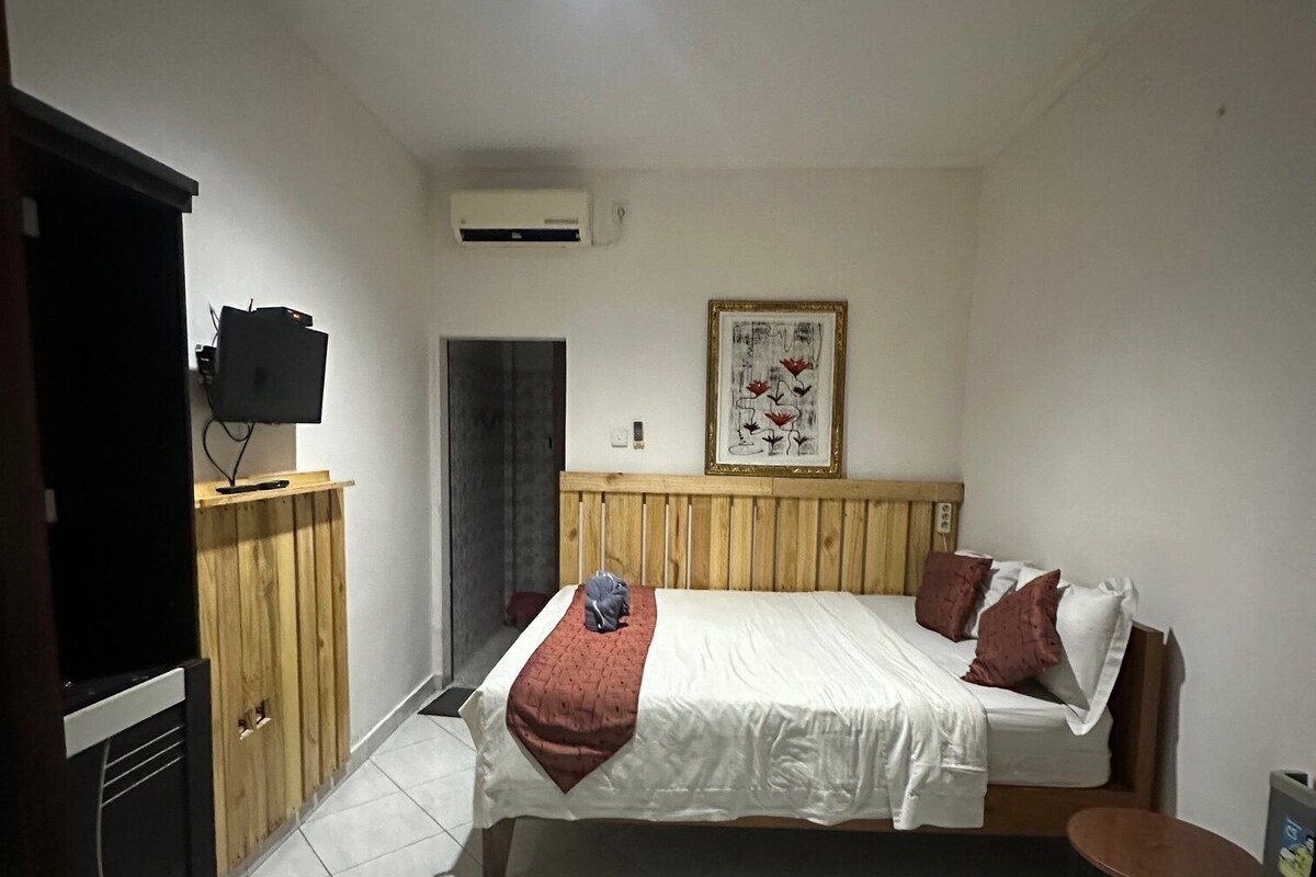 A bedroom with a bed, a television on a wooden cabinet, a small table, and a framed picture on the wall.