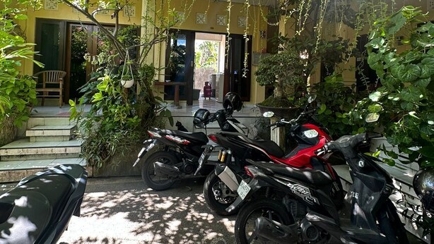 Un patio con dos motocicletas estacionadas bajo árboles, la entrada de un edificio y escalones que conducen a una puerta.
