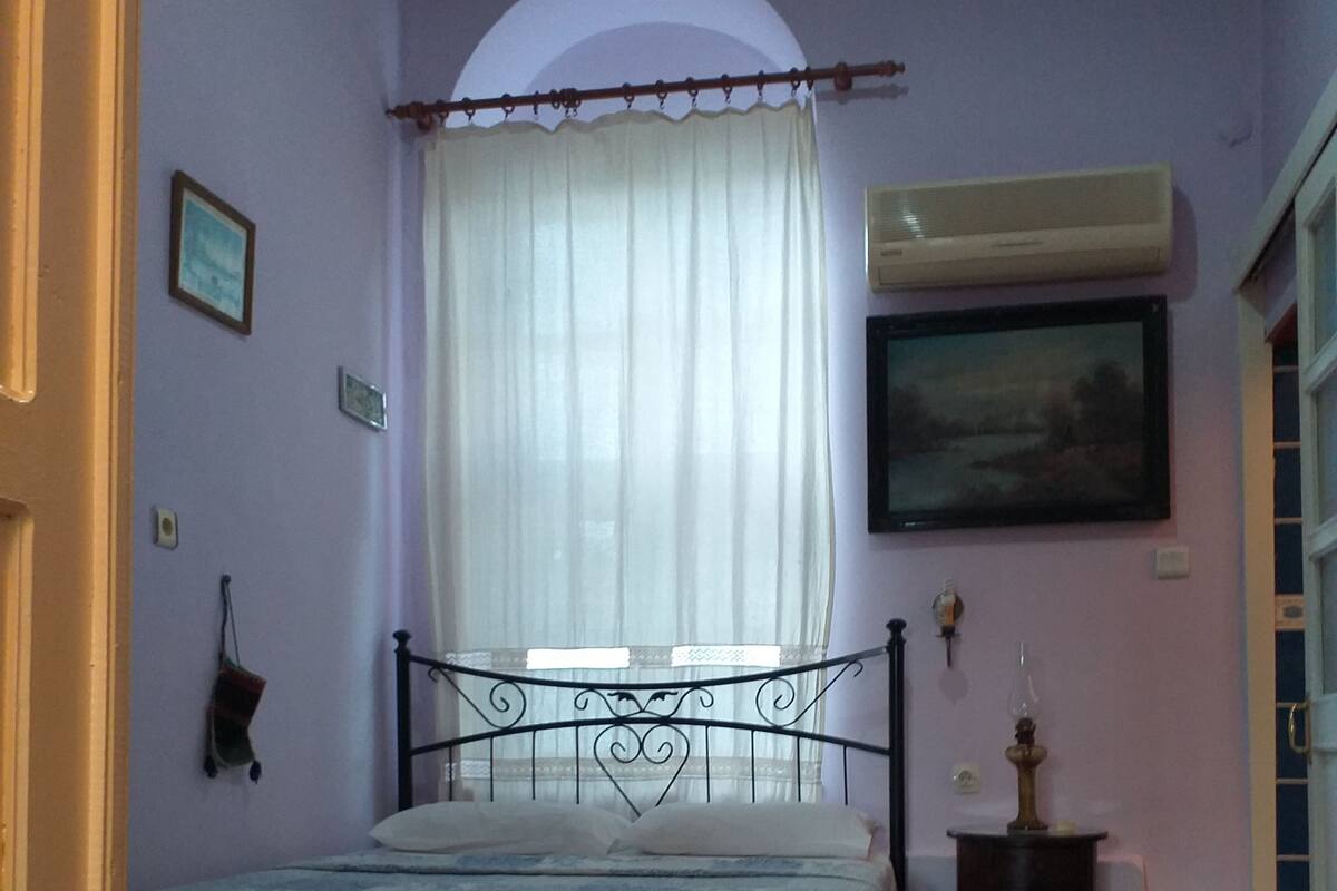 Een slaapkamer met een bed, een stoel, een klein tafeltje, een wandairco en een schilderij.