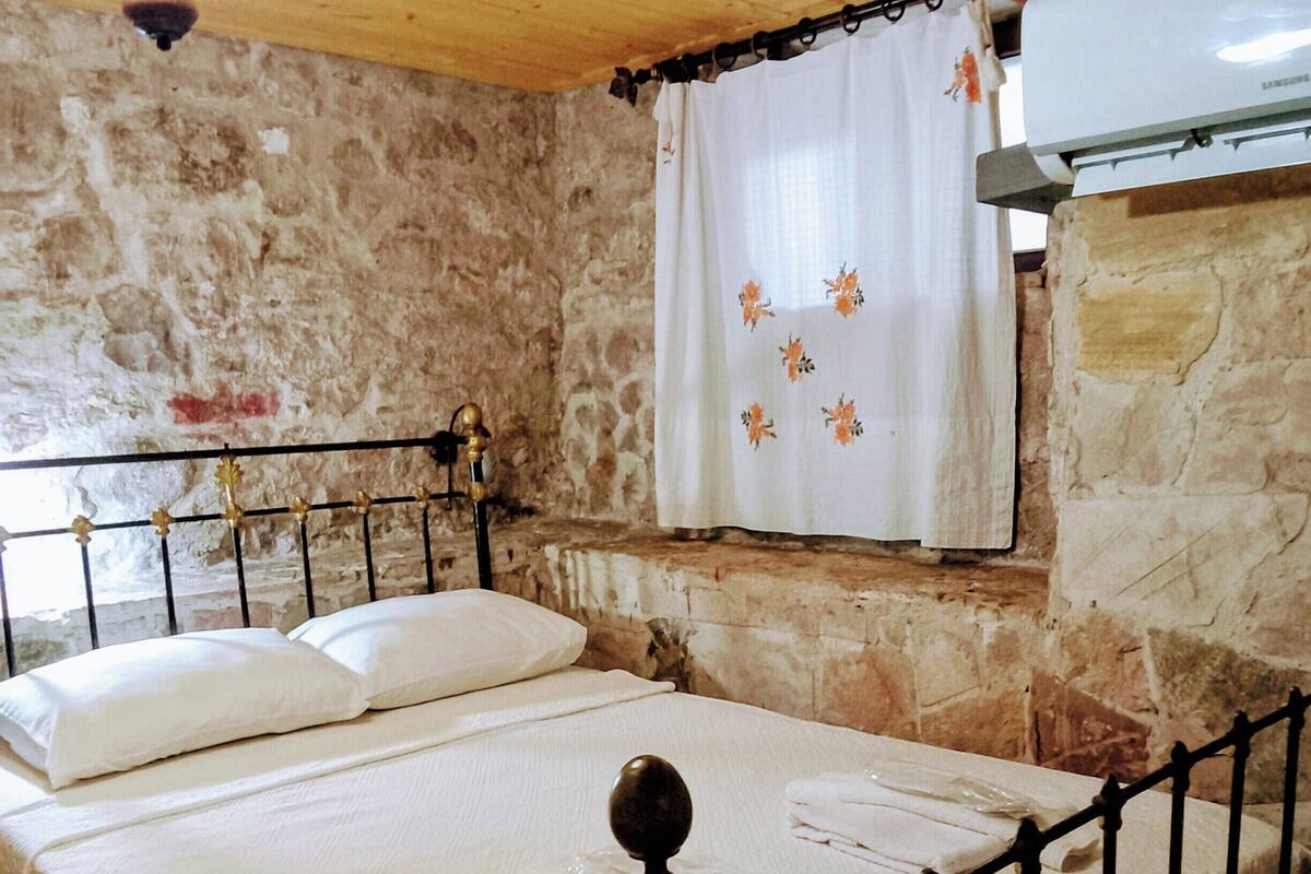 Een kamer met een stenen muur, een houten plafond, een bed met metalen frame en een hangende lamp.