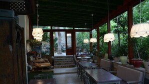 Un café al aire libre con mesas y sillas de madera, plantas en macetas y lámparas colgantes.
