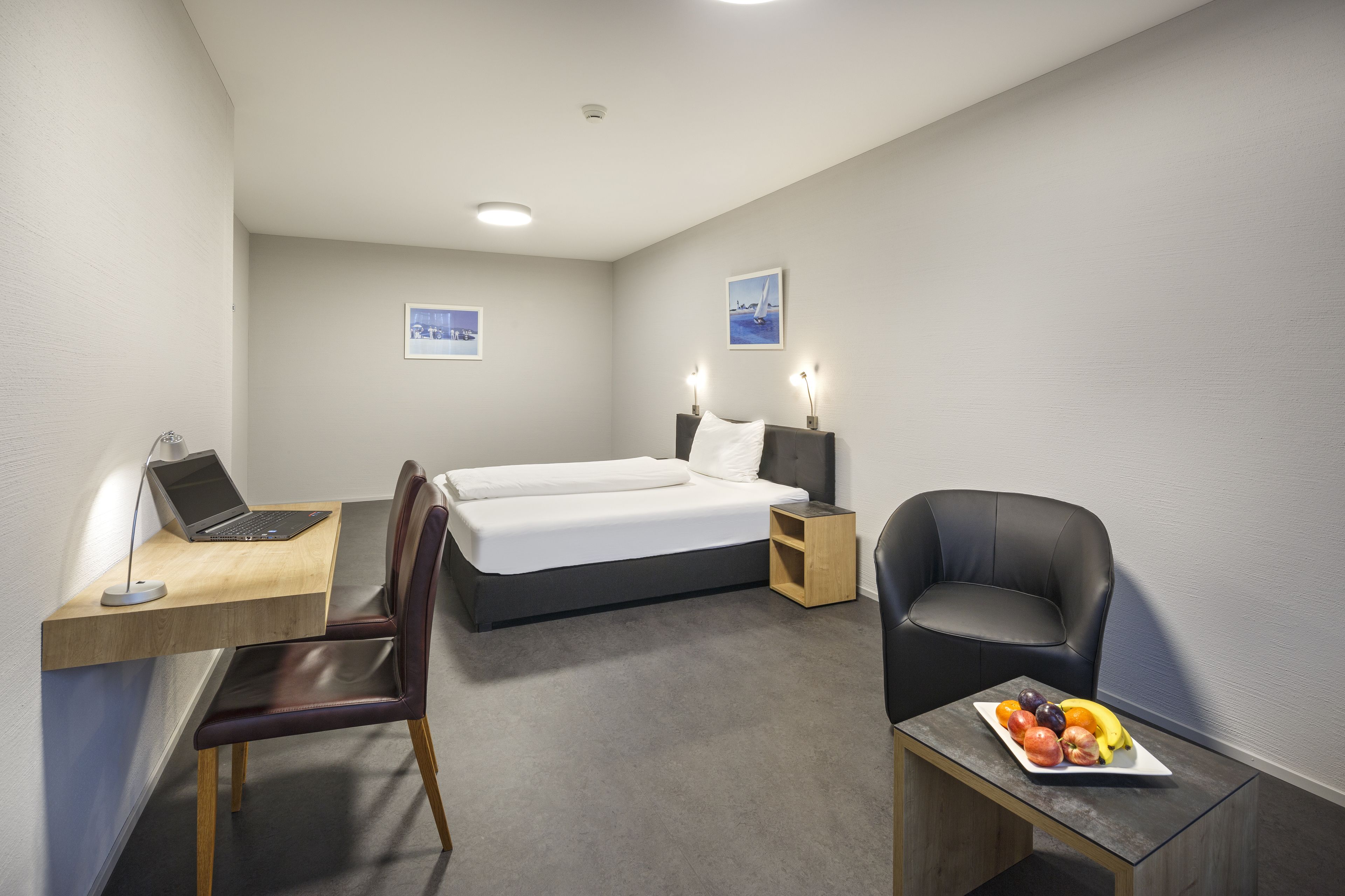 Habitación de hotel con cama, escritorio con una computadora portátil, silla, pequeña mesa con fruta y dos lámparas de pared.
