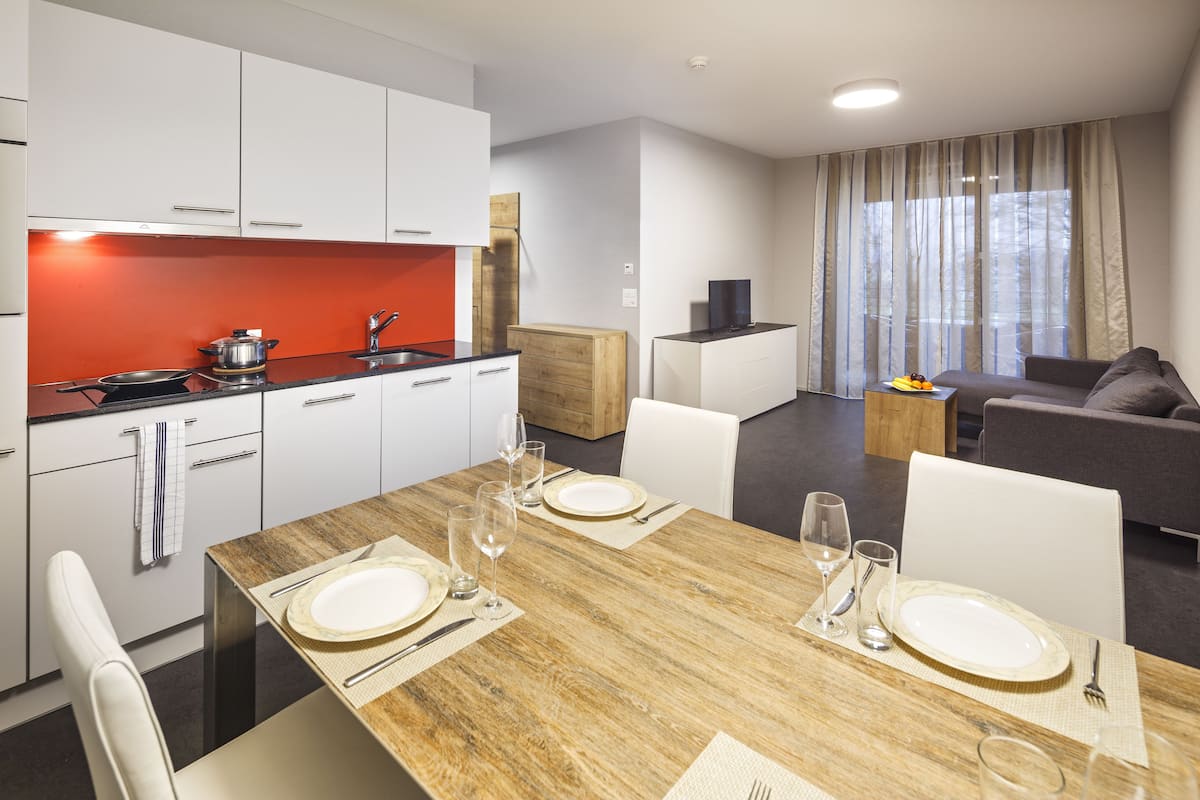 A modern kitchen with white cabinets, a wooden table set for four, and a dining area with a grey sofa and a TV.