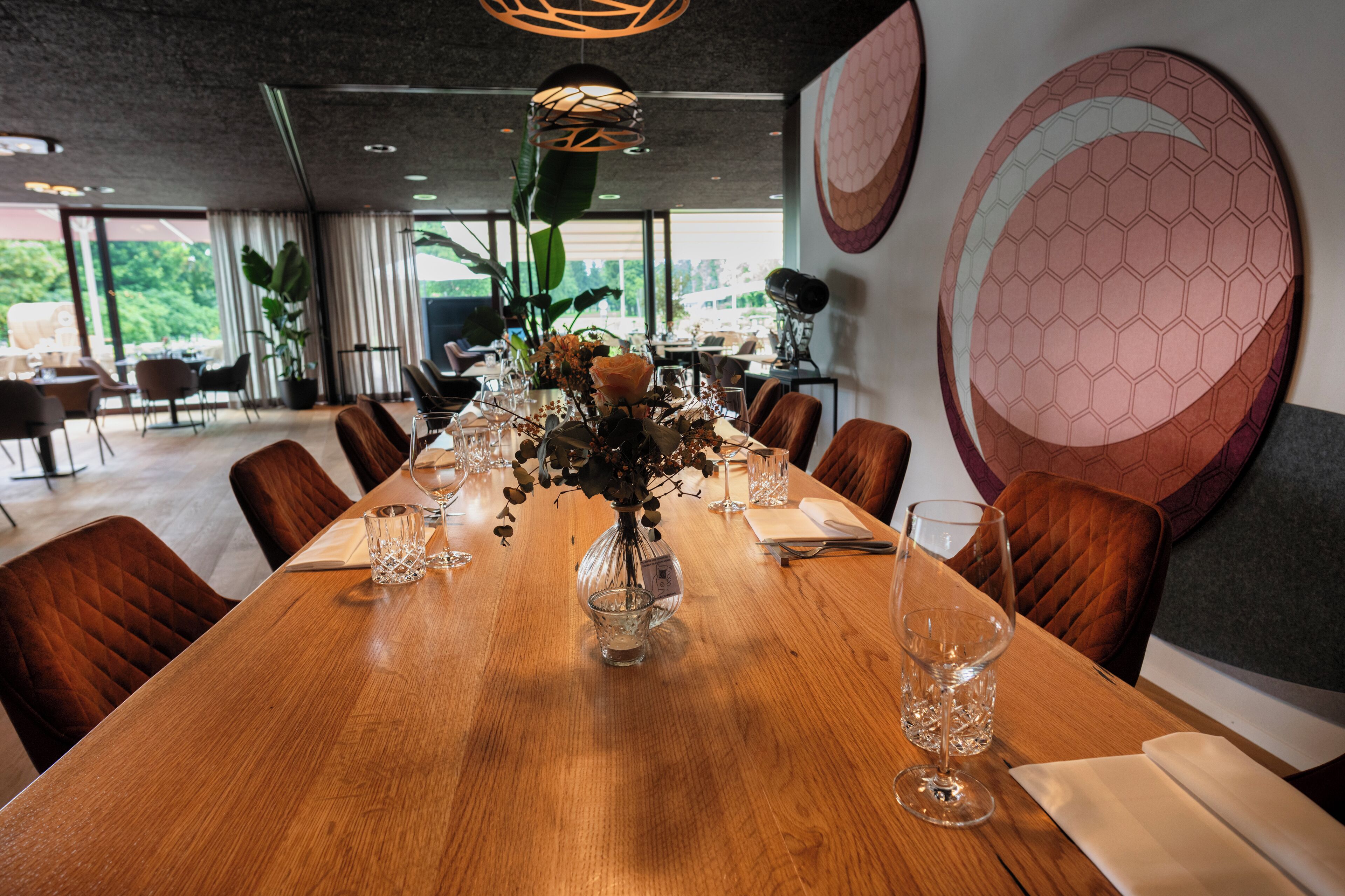 A long wooden dining table set with glassware and a vase of flowers, surrounded by brown upholstered chairs.