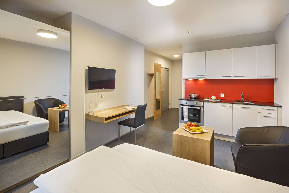 A hotel room with a bed, desk, chair, kitchenette, and a TV mounted on the wall.