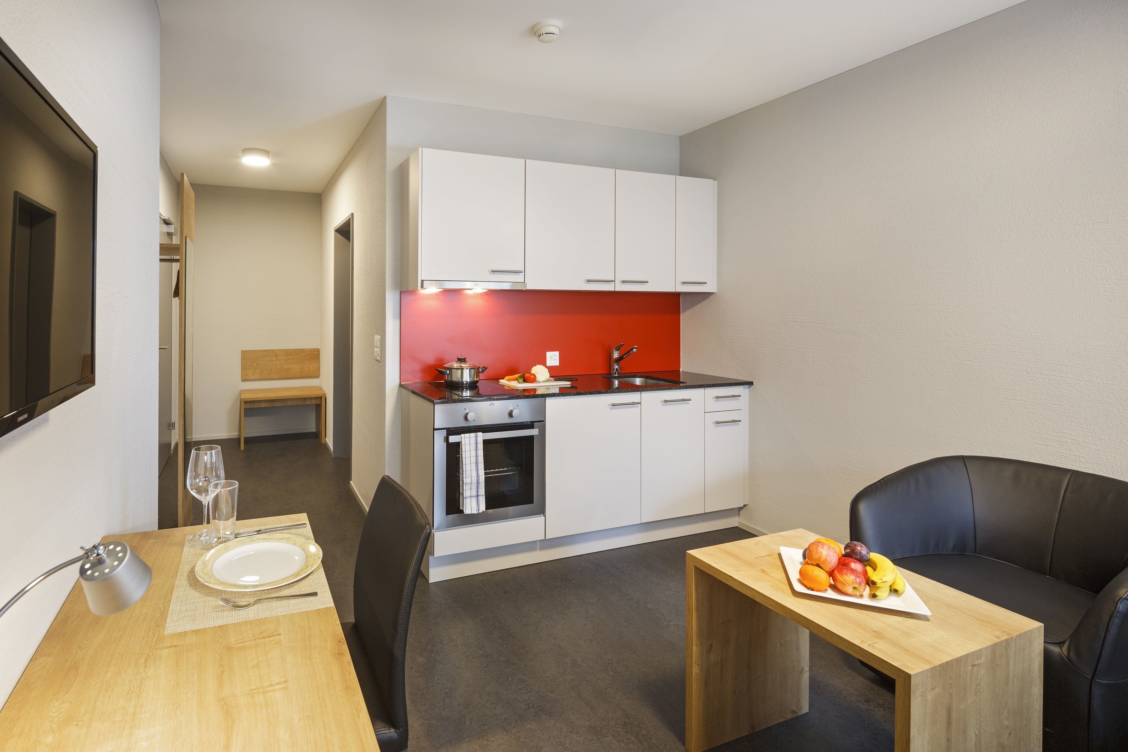 Una cocina compacta con armarios blancos, salpicadero rojo y una zona de comedor con una mesa dispuesta para dos.