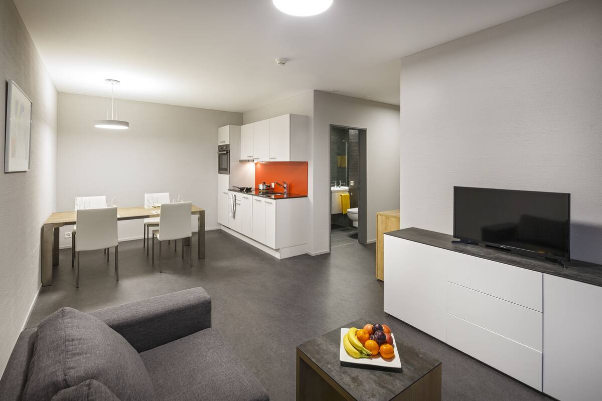A modern living room with a gray sofa, a coffee table with fruit, a flat-screen TV, and a kitchen area with white cabinets and a red backsplash.