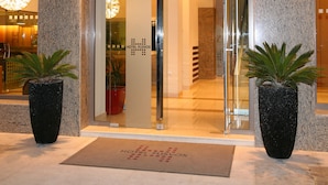The entrance to Hotel Fisvos with a glass door, a doormat with the hotel's name, and two potted plants on either side. - Hotel Flisvos (Kalamata)