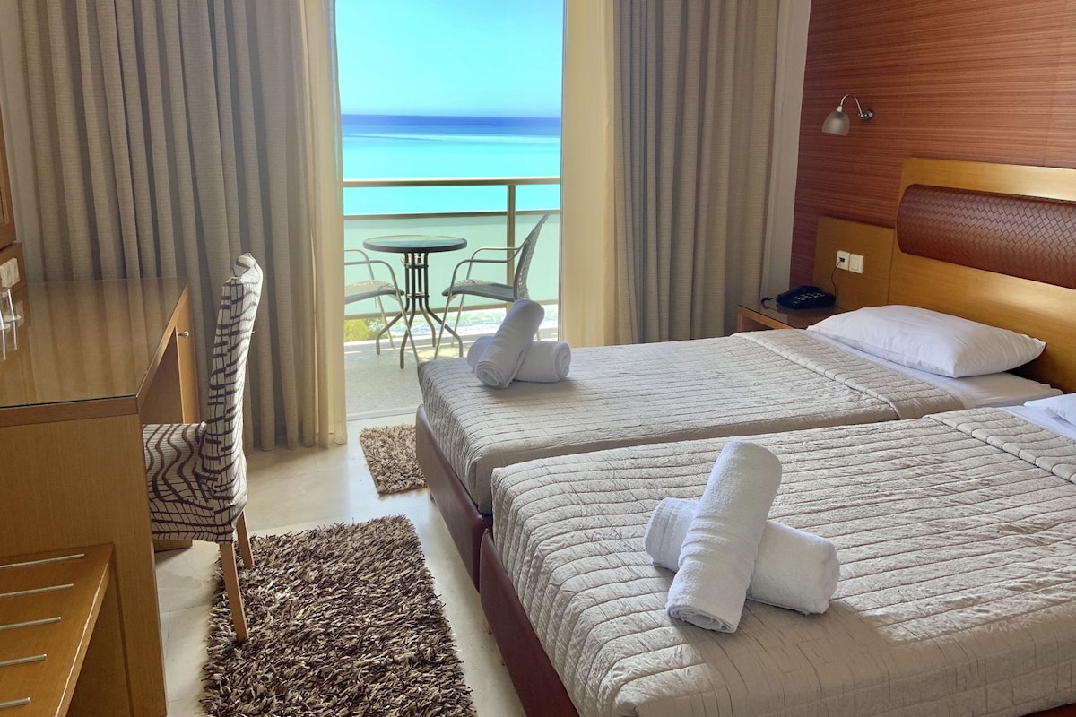 Habitación de hotel con dos camas, un escritorio, una silla, una mesita y un balcón con vista al mar.